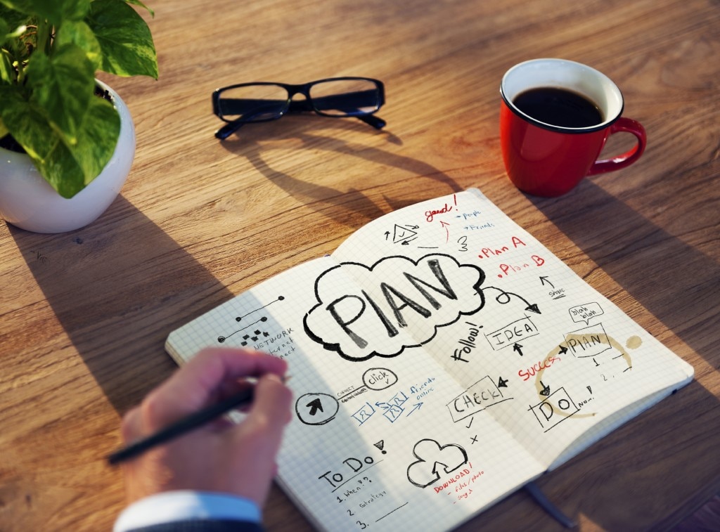 Escribir a mano en cuaderno con planos en un escritorio con una planta, vasos y una taza de café