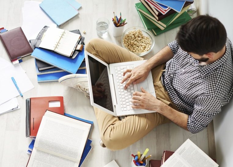 Estudiante sentado en el suelo con un portátil en su regazo rodeado de cuadernos, libros y papeles