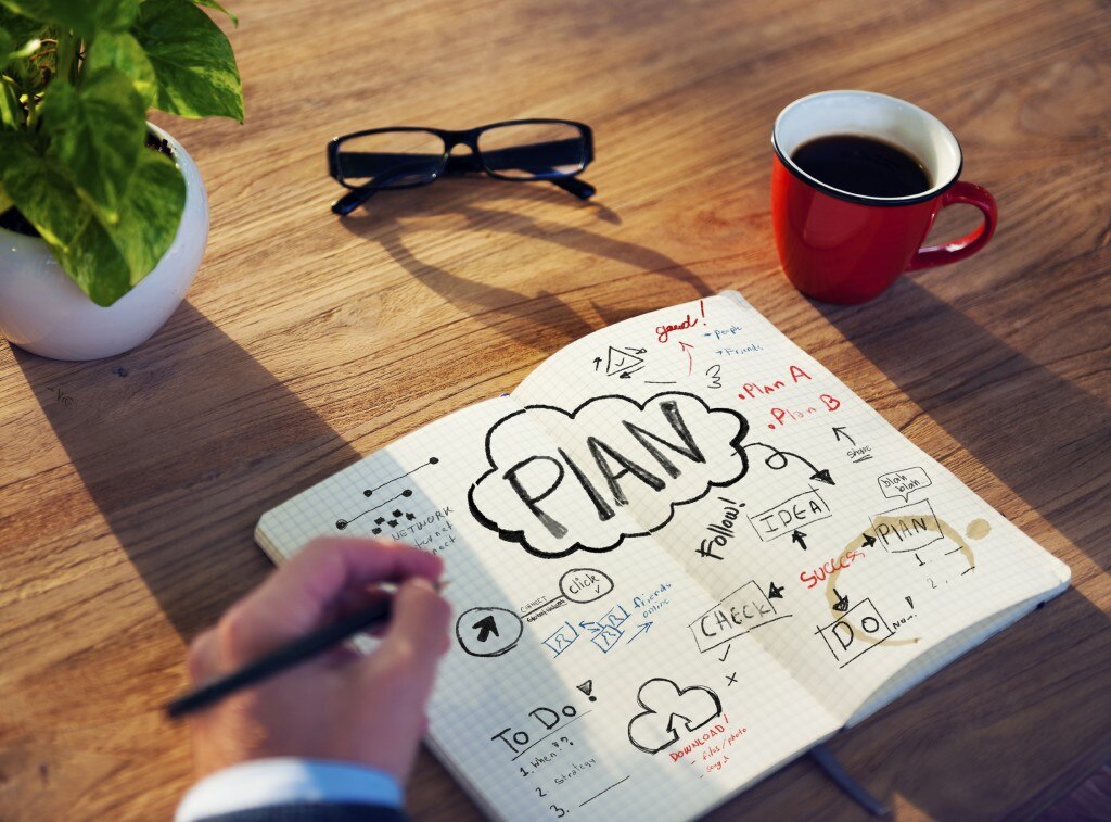 Escribir a mano en cuaderno con planos en un escritorio con una planta, vasos y una taza de café
