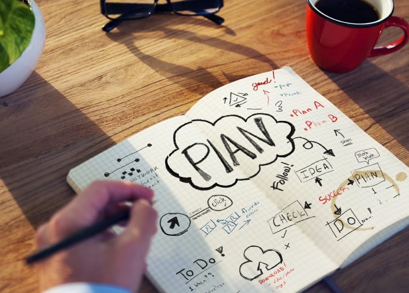 Escribir a mano en cuaderno con planos en un escritorio con una planta, vasos y una taza de café